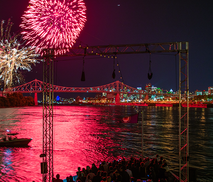 A vibrant fireworks display in a black sky above the AML Cavalier Maxim ship in Montreal, with the city illuminated in the distance.