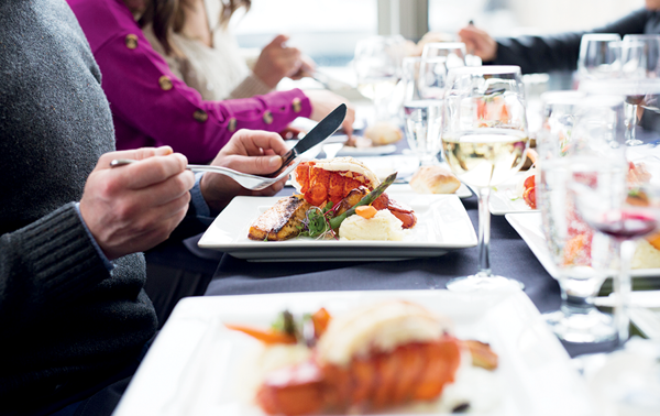 Gros plan sur des gens qui dînent avec une assiette de homard au premier plan accompagnée de verres de vin et d'autres plats autour. 
