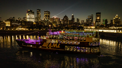 Vue aérienne du navire AML Cavalier Maxim entrant dans le Vieux-Port de Montréal de nuit avec vue sur la skyline de la ville en arrière plan.