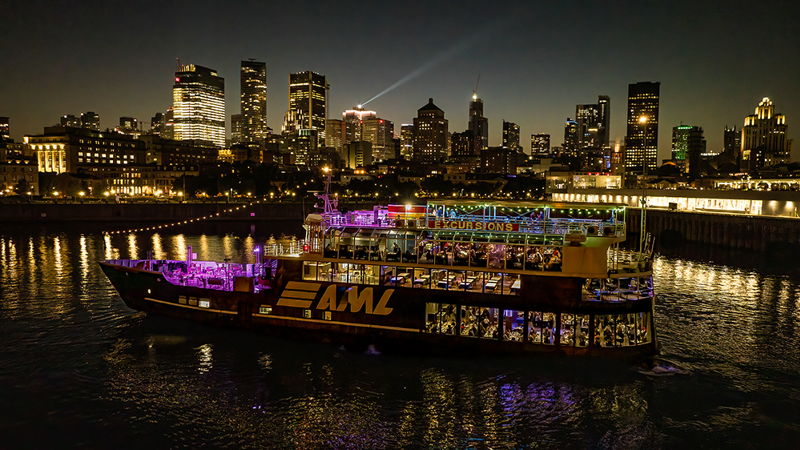 Vue aérienne du navire AML Cavalier Maxim entrant dans le Vieux-Port de Montréal de nuit avec vue sur la skyline de la ville en arrière plan.
