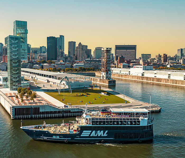 Vue aérienne du navire AML Cavalier Maxim stationné devant la Tour du Port de Montréal de soir avec vue sur la skyline de la ville et le coucher de soleil en arrière plan.