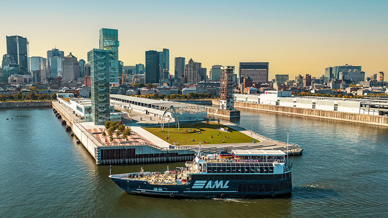 Vue aérienne du navire AML Cavalier Maxim stationné devant la Tour du Port de Montréal de soir avec vue sur la skyline de la ville et le coucher de soleil en arrière plan.