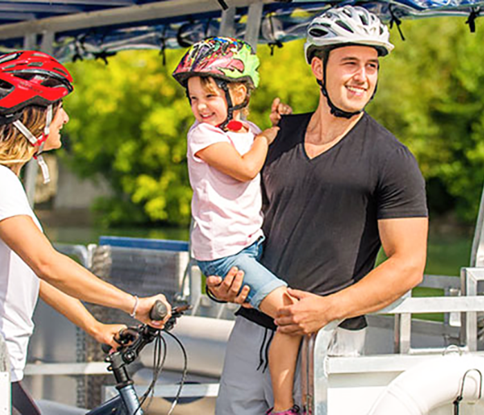 A couple and a child enjoying a ride on a river shuttle on the St. Lawrence River in Montreal. The woman is holding a bicycle and wearing a helmet, while the man is carrying the child in his arms.