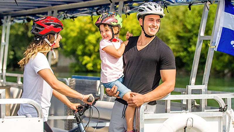 A couple and a child enjoying a ride on a river shuttle on the St. Lawrence River in Montreal. The woman is holding a bicycle and wearing a helmet, while the man is carrying the child in his arms.