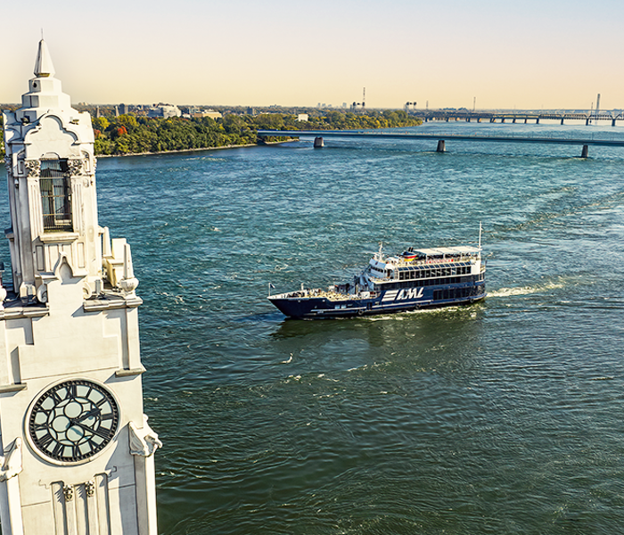 Le navire AML Cavalier Maxim naviguant sur le fleuve Saint-Laurent avec au premier plan la tour de l'horloge de Montréal et en arrière plan l'Ile Sainte Hélène et le Pont Samuel de Champlain avec une lumière dorée.