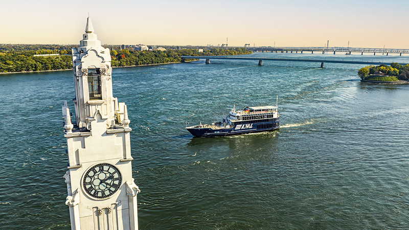 Le navire AML Cavalier Maxim naviguant sur le fleuve Saint-Laurent avec au premier plan la tour de l'horloge de Montréal et en arrière plan l'Ile Sainte Hélène et le Pont Samuel de Champlain avec une lumière dorée.