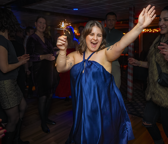 Une femme vêtue d'une robe bleue tenant joyeusement un feu de bengale en l'air célébrant Noël entourée d'amis.