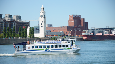 A river shuttle sailing on the St. Lawrence River, with the river in the foreground and the Clock Tower and the Molson Brewery building in the background.