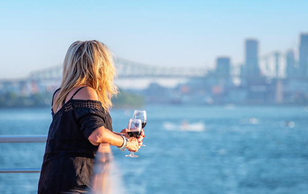 Une femme tient un verre de vin rouge sur l'une des terrasses du navire AML Cavalier Maxim naviguant sur le fleuve Saint-Laurent avec une vue sur la skyline de Montréal en arrière plan. 