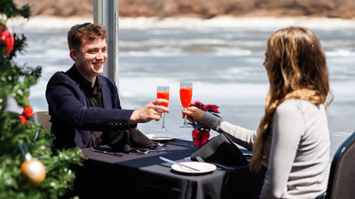 Couple complice et souriant en train de trinquer à table avec un cocktail lors d'une croisière brunch de Noël avec un sapin de Noël décoré au premier plan à gauche