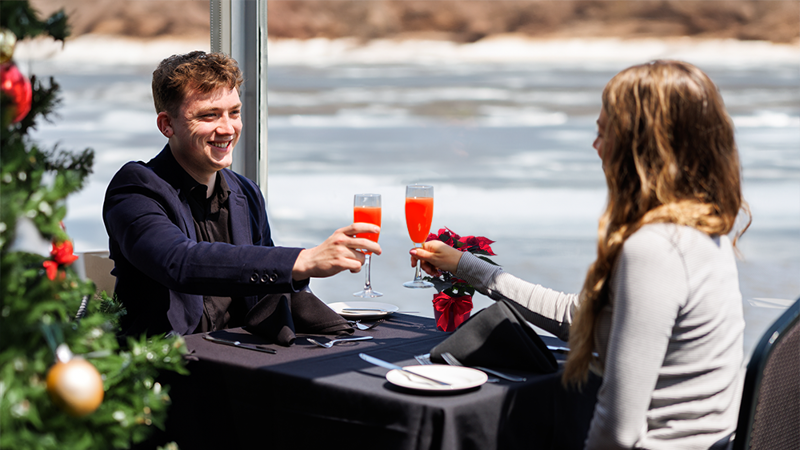 Couple complice et souriant en train de trinquer à table avec un cocktail lors d'une croisière brunch de Noël avec un sapin de Noël décoré au premier plan à gauche