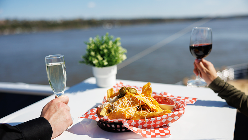 Deux personnes attablées avec vue sur le fleuve, l'une tenant un verre de vin et l'autre une flûte de champagne, partageant un panier de nachos. 