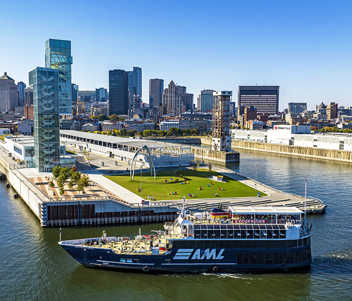 Vue aérienne du navire AML Cavalier Maxim stationné devant la Tour du Port de Montréal de jour avec vue sur la skyline ensoleillée de la ville en arrière plan.