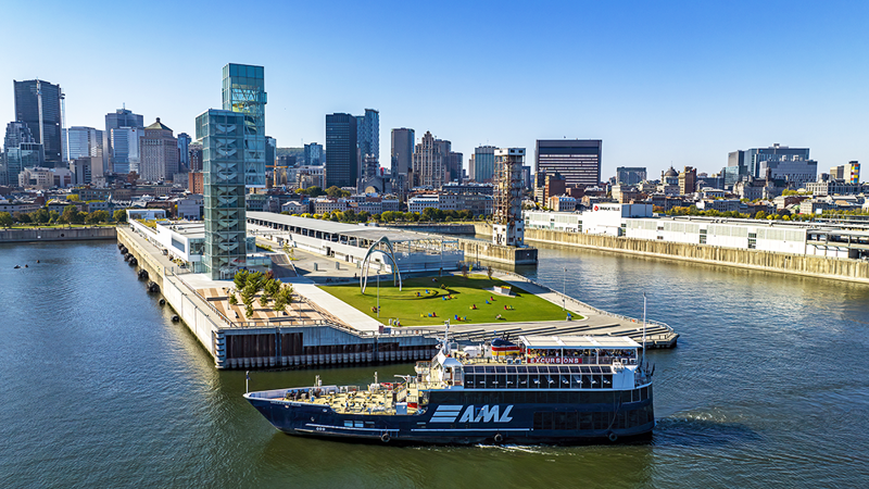 Vue aérienne du navire AML Cavalier Maxim stationné devant la Tour du Port de Montréal de jour avec vue sur la skyline ensoleillée de la ville en arrière plan.