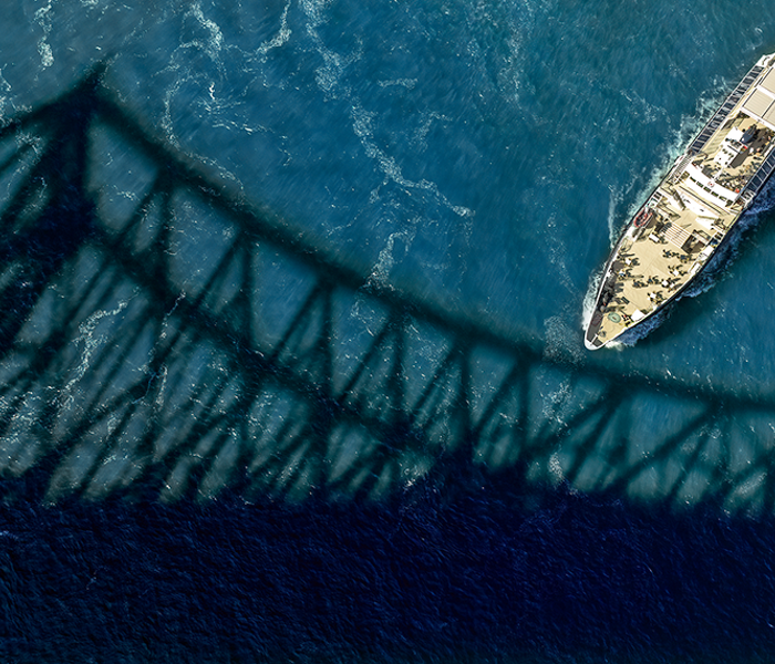Vue aérienne du navire Cavalier Maxime naviguant sur l'étincelant fleuve Saint-Laurent avec l'ombre du Pont Jacques Cartier au premier plan. 
