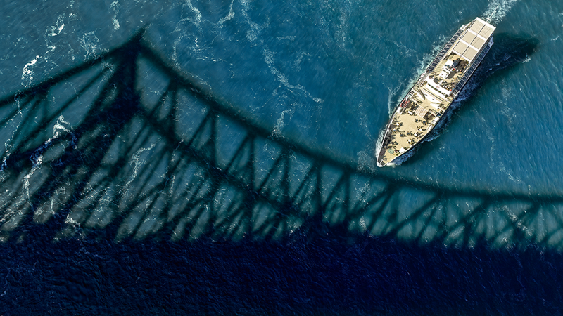 Vue aérienne du navire Cavalier Maxime naviguant sur l'étincelant fleuve Saint-Laurent avec l'ombre du Pont Jacques Cartier au premier plan. 