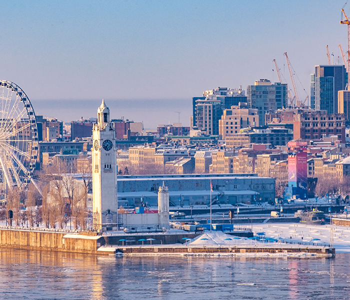 Vue de Montréal en hiver depuis le Vieux-Port avec la Tour de l'Horloge au premier plan, puis la grande roue derrière, et la skyline de Montréal enneigée en arrière plan.