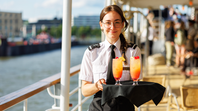Serveuse tenant un plateau avec des cocktails orangés sur la terrasse extérieure du navire AMl Cavalier Maxim à Montréal. 