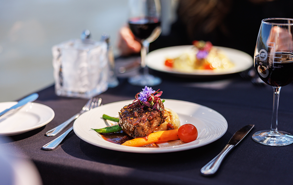 Un plat de steak gourmet garni d'une fleur, accompagné d'un verre de vin rouge, avec vue sur le cadre serein d'une vue sur l'eau.