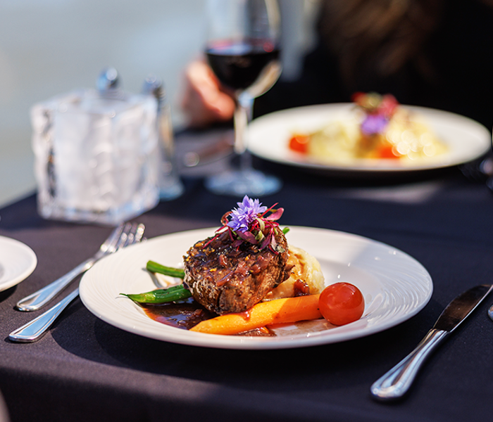 Un plat de steak gourmet garni d'une fleur, accompagné d'un verre de vin rouge, avec vue sur le cadre serein d'une vue sur l'eau.