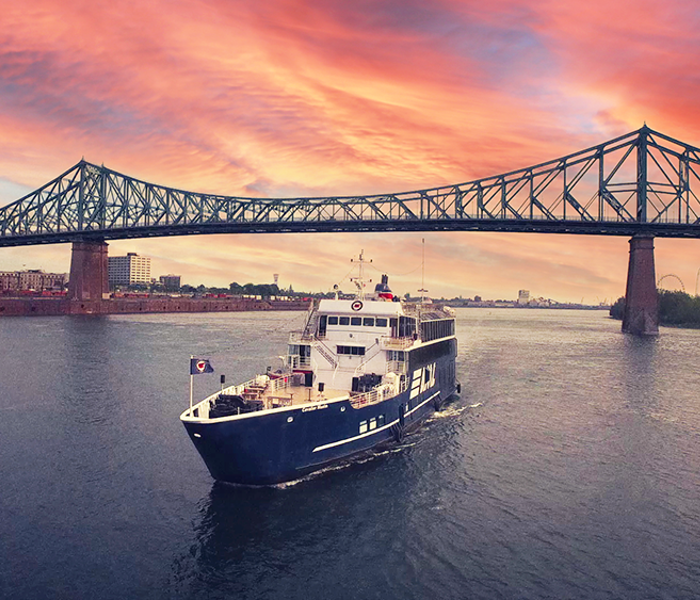 Le navire AML Cavalier Maxim naviguant seul sur le fleuve Saint-Laurent avec le Pont Jacques Cartier et un coucher de soleil vibrant en arrière plan.