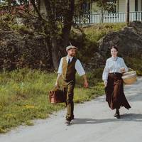 Deux personnages habillés en tenue d'époque marche sur Grosse Île. Ils descendent une petite pente. Il y a une maison d'époque en arrière plan 