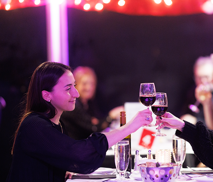 Couple souriant et complice assis à une table trinquant avec un verre de vin rouge avec des décorations de Noël en arrière plan